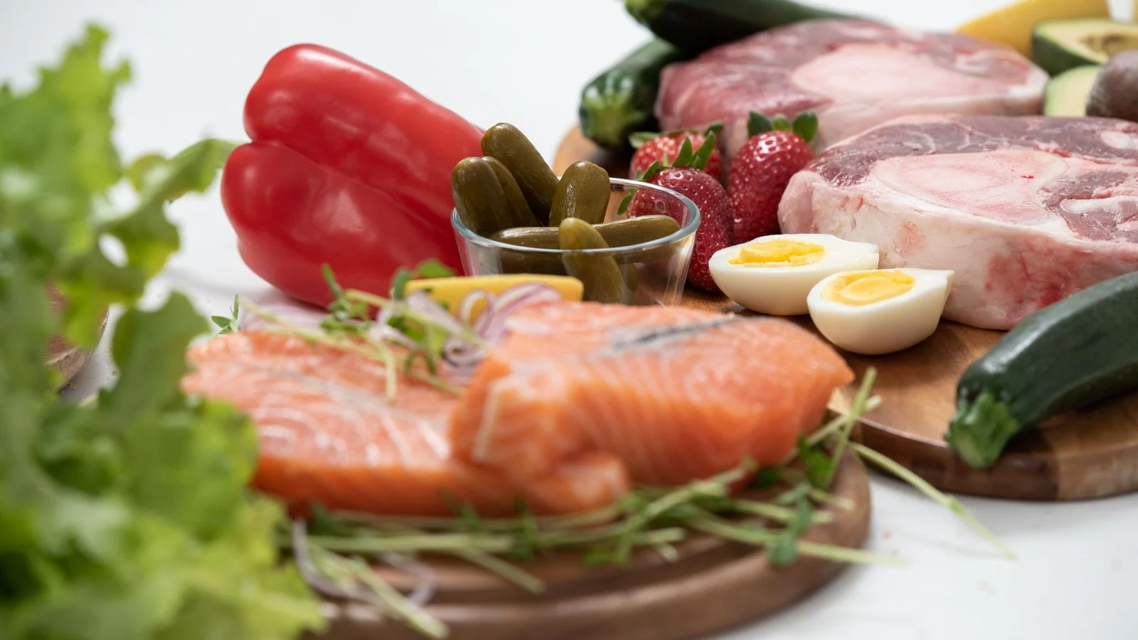 Ketoproducten op houten planken: zalm, rauw vlees, gekookte eieren, paprika, augurk en sla