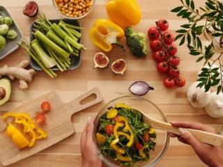 Houten tafel met verse groente, kikkererwten, vijgen en avocado terwijl iemand een salade mengt voor een mediterraan dieet