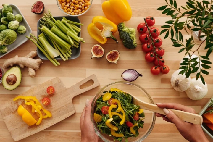 Houten tafel met verse groente, kikkererwten, vijgen en avocado terwijl iemand een salade mengt voor een mediterraan dieet