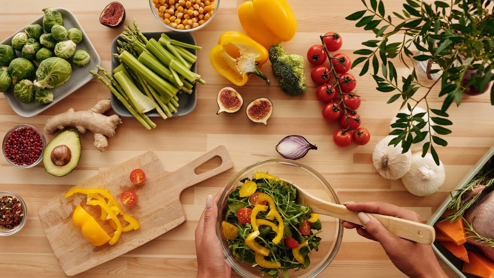 Houten tafel met verse groente, kikkererwten, vijgen en avocado terwijl iemand een salade mengt voor een mediterraan dieet